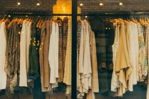 A clothing store display with neatly hung garments on wooden hangers. The collection includes sweaters, jackets, and shirts in neutral tones like beige, brown, and cream, illuminated by warm lighting.