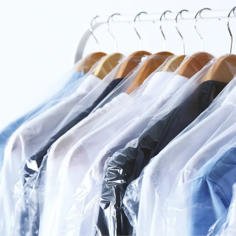 A row of shirts in various colors, including white, blue, and black, hang on wooden hangers. Each shirt is covered with a clear plastic garment bag, all suspended from a metal clothing rack.