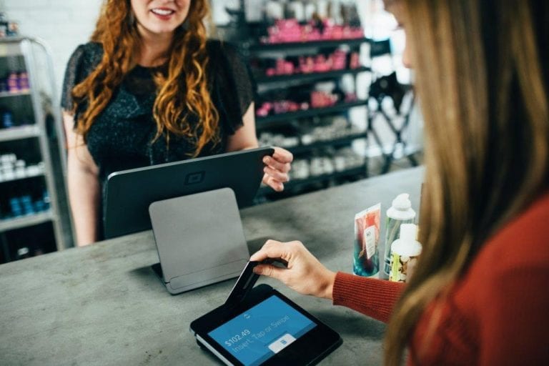 A woman with long red hair stands behind a counter, smiling at a customer using a card reader. The customer holds a credit card over the payment terminal. Shelves with various products in the background suggest a retail environment.