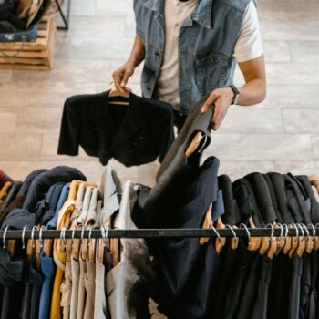 A person browses through jackets on a clothing rack, holding two blazers—one in each hand, suspended gracefully on Longlife Plastic Hangers. The wooden floor adds warmth while other clothes and a stack of crates form the backdrop.