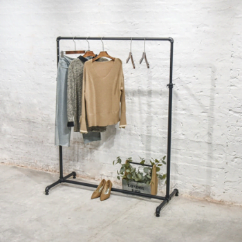 A minimalist clothing rack against a white brick wall holds a beige sweater, a patterned shirt, and light blue pants. Two empty hangers are beside them. Below, a pair of brown heels and a box with a plant add decoration.