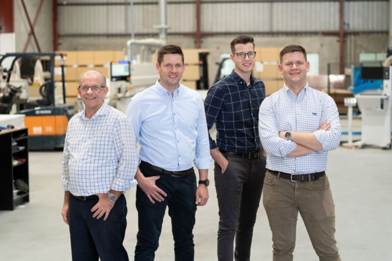 Four men stand in a warehouse, smiling at the camera. Dressed in casual business attire, they embody the essence of quality shopfitting. Behind them, industrial equipment and boxes hint at their expertise in supplying retailers large and small, all under a high ceiling with exposed beams.