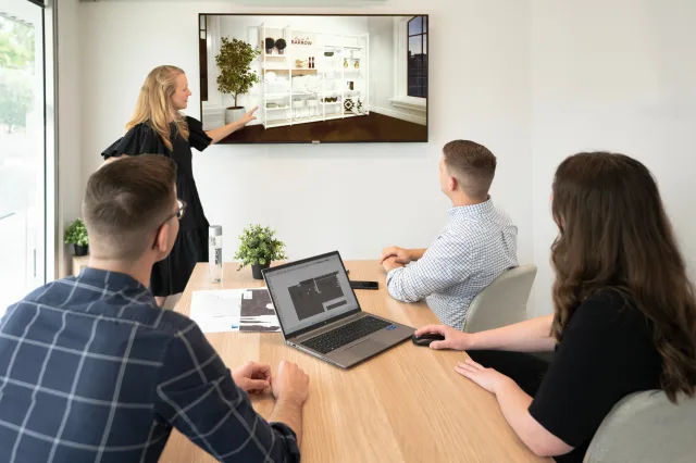 A woman stands pointing at a wall-mounted screen displaying a modern room interior design, while three colleagues, two men and one woman, seated at a table with laptops, attentively watch. The room's neutral tones reflect quality shopfitting ideal for retailers large and small.