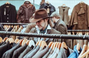 man in brown cowboy hat in front of Clothing Racks with hanging suit jackets looking to purchase. Mannequins in Background. 