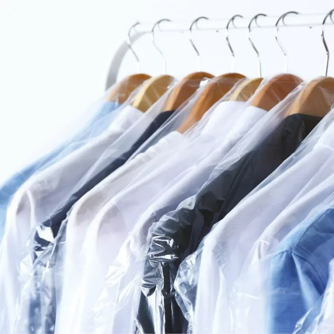 A collection of shirt-length dresses, neatly organized in Dress Length Garment Covers, hangs on wooden hangers to display a diverse array of colors—white, blue, and black. These sharp garments are elegantly showcased on a clothing rack set against a light backdrop.