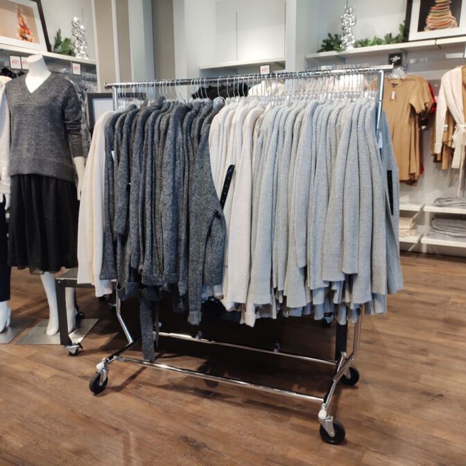 A clothing store display showcases a Single Rail Travellers Rack Chrome with gray and beige sweaters. A nearby mannequin models a gray sweater with a black skirt, while the neatly folded clothes on shelves in the background accentuate the warm wood-textured floor.