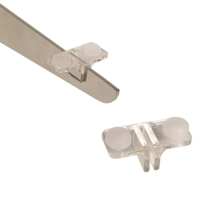 A clear plastic clip is being attached to a slim metallic tool, while another clip lies nearby. The Double Sided Shelf Clips are equipped with small, round white pads on each end, providing a secure grip and versatile use.