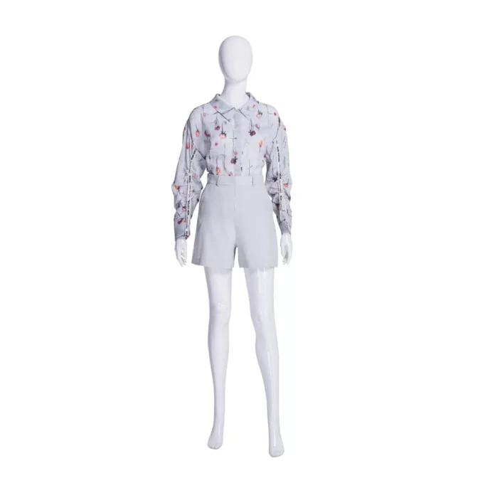 A Patsy Pose 1 Plastic Gloss White Female Mannequin displays a long-sleeve blouse featuring abstract red and gray designs, along with light-colored high-waisted shorts. The mannequin’s glossy finish shines brilliantly against the plain white backdrop.