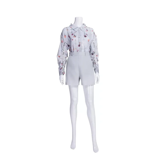 A Patsy Pose 1 Plastic Gloss White Female Mannequin is elegantly dressed in a floral patterned long-sleeve blouse and high-waisted light gray shorts, set against a gloss white backdrop to enhance the chic and sophisticated style.