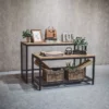 A stylish room features the Industrial Display Table Set, showcasing two stacked wooden tables framed in metal. The top table is adorned with plants, a framed picture, books, and decor, while the lower table holds baskets with rolled towels and small potted plants against a textured gray wall.
