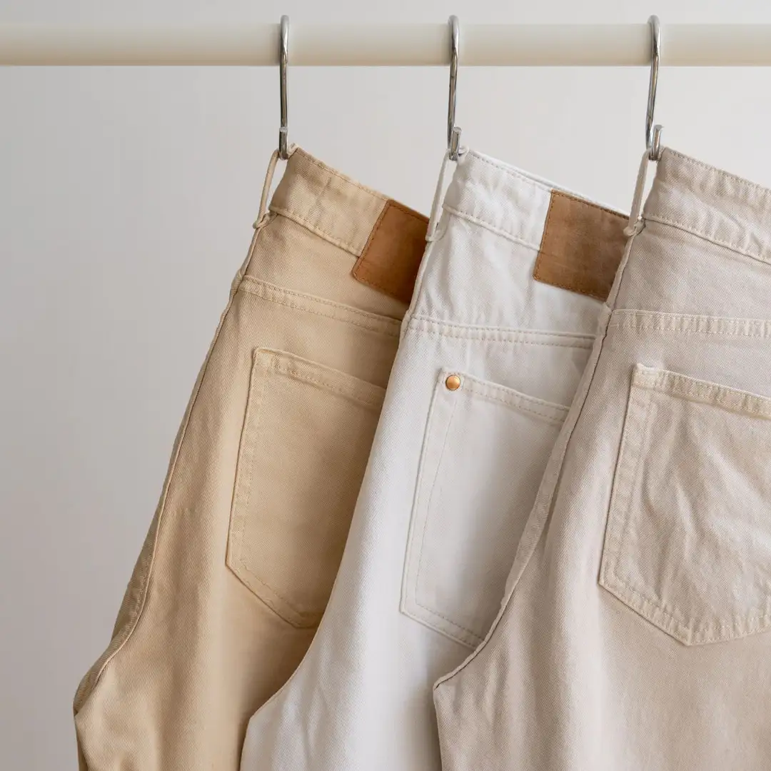 Three pairs of jeans in beige, white, and light khaki hang on hooks against a neutral background. Each product features visible back pockets and belt loops, showcasing a casual and minimalist style. This range highlights simplicity and versatility for every wardrobe.