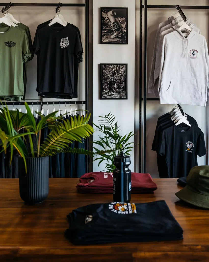 T-shirts and hoodies hang on racks above folded shirts on a wooden table with green plants, a water bottle, and a hat. Framed black-and-white photos are on the wall, showcasing one of the Featured Projects Apex Display is known for.