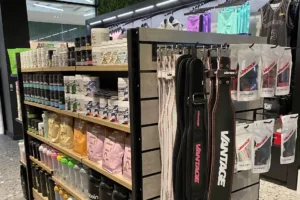 The East Coast Supplements display features shelves brimming with fitness essentials like protein powders, shaker bottles, and exercise accessories. In the foreground, weightlifting belts hang on a rack amidst vibrant packaging and trendy fitness apparel.