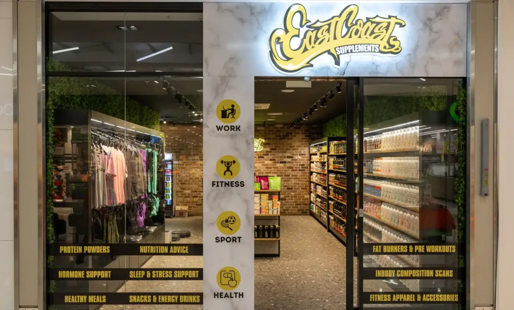 Storefront of East Coast Supplements, with glass doors, yellow logo, and icons for work, fitness, sport, and health. Inside are shelves of supplements, apparel, and health products—showcasing a Featured Projects Apex Display—with clothing displayed on the left.