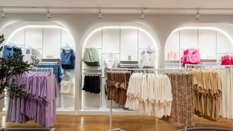 A clothing store interior showcases neatly arranged racks of colorful garments, highlighting brand partnerships with tops and dresses in shades of purple, white, brown, blue, and pink. The space is well-lit with wooden flooring and adorned with plants for a touch of natural decor.