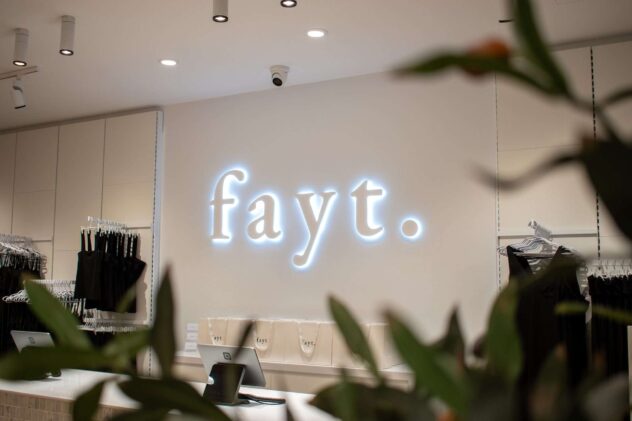 A clothing store interior with fayt. illuminated on the wall. Dark clothing items hang on the left. The checkout counter is visible, with shopping bags displayed in the background. Green leaves partially frame the scene.