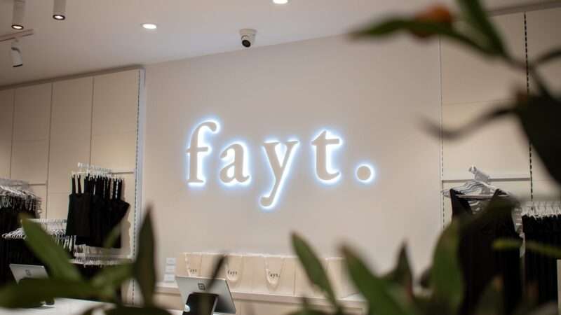 A clothing store interior with fayt. illuminated on the wall. Dark clothing items hang on the left. The checkout counter is visible, with shopping bags displayed in the background. Green leaves partially frame the scene.