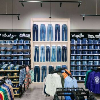 A clothing store display featuring shelves stocked with folded jeans and shirts. Various brands like Levis, Wrangler, and Sportscraft are visible on signs. In the center, several pairs of jeans are showcased vertically on a lit wall panel.