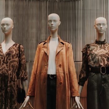 Three mannequins strategically placed in the retail display showcase autumn outfits: a paisley dress, a beige trench coat with a white top, and a patterned blouse with a denim skirt. They stand against a textured, vertical-striped background.