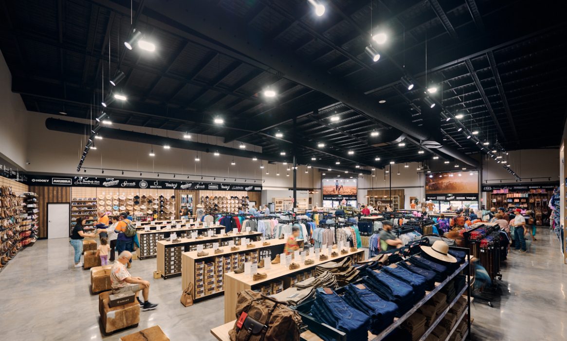 A spacious, well-lit clothing store with rows of wooden shelves displaying folded apparel invites customers to browse. The ceiling has exposed beams, and large lights hang overhead. A wall showcases hats, with bags in the foreground—a perfect reflection of W. Titley & Cos timeless style.